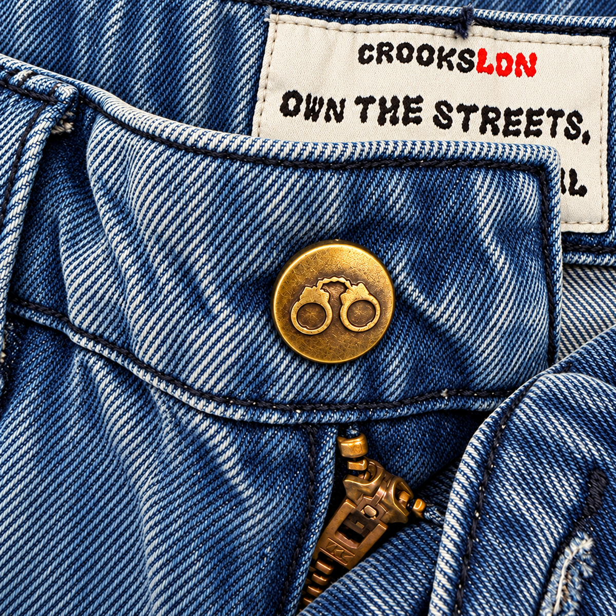 CROOKSLDN Blue Wash Yard Jeans — antique brass handcuff button, zipper and inside OWN THE STREETS label.